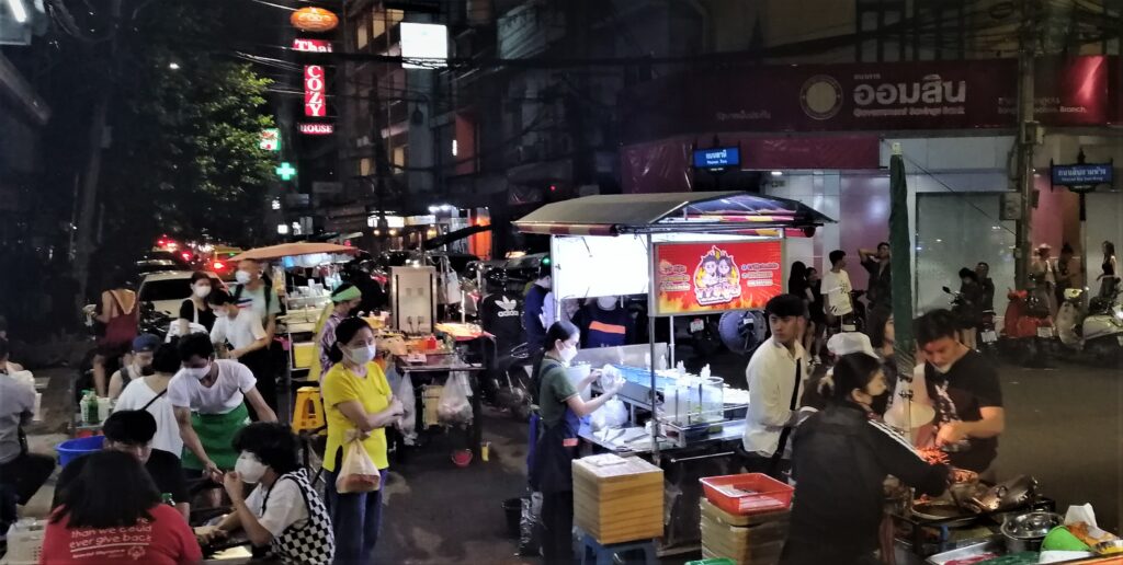 Lots of people and some food stalls outside in the urban night.  From the post: Wiped Out After A Journey Across the Earth.