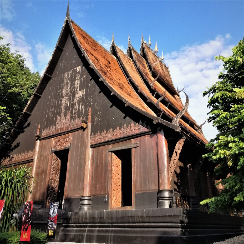 Brown rustic building design with four very steep roofs visible under a billowy and blue sky. In the post: Temple Drifting and Coffee Sipping in Chiang Rai.