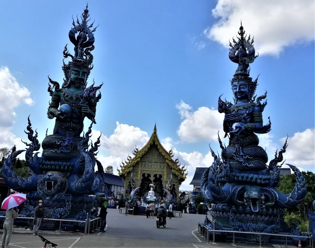 Two ginormous blue mythical statues of Gods under blue and cloudy sky with temple in the background. In the post: Temple Drifting and Coffee Sipping in Chiang Rai.
