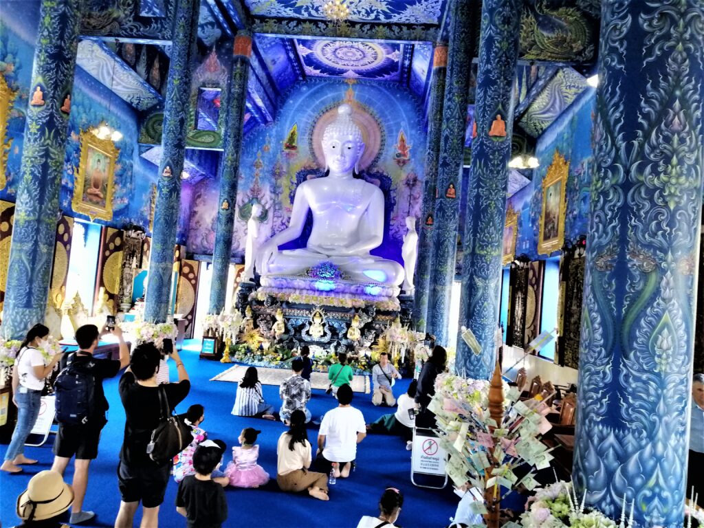 A huge Buddha and worshipers and tourists inside a magically religious building.  In the post: Temple Drifting and Coffee Sipping in Chiang Rai.