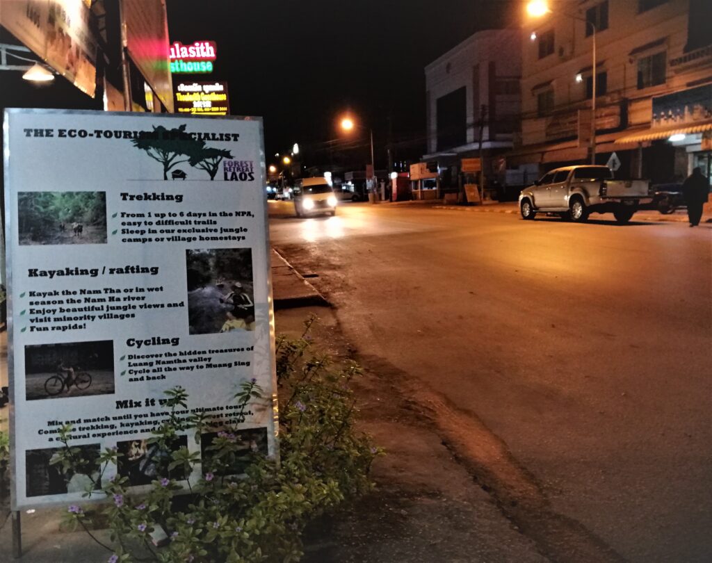 A tourist van and pick up truck on the main road in the evening in northern Laos.  And an eco tourism activities sign. From the post: Luang Namtha: Why I Spent a Week in a Virtual Ghost Town.