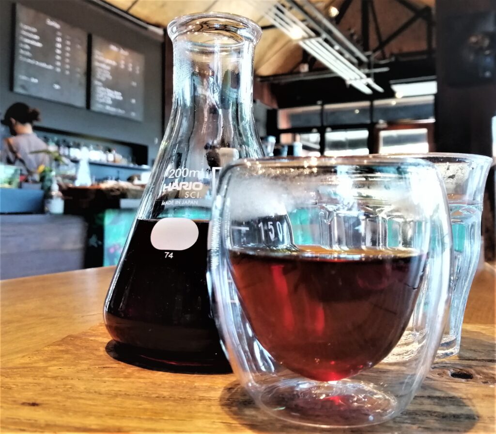 A V60 drip coffee sits in a Hario decanter (carafe) behind a sleekly curved class that some coffee has been poured into.  In the post: Temple Drifting and Coffee Sipping in Chiang Rai.