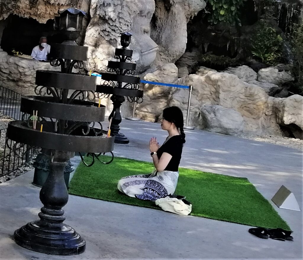 A woman prays in a kneeling position outside on a green mat. In the post: Temple Drifting and Coffee Sipping in Chiang Rai.