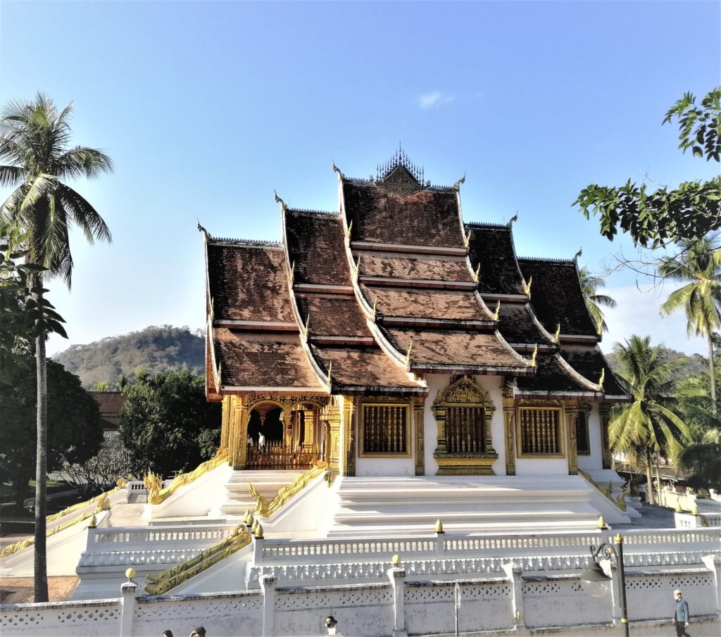 A stunning rustic-roofed temple enclosed by white walls and surrounded by lushness under a wide-open blue sky. In the post: Infinite Earth Art: Temples of Luang Prabang.