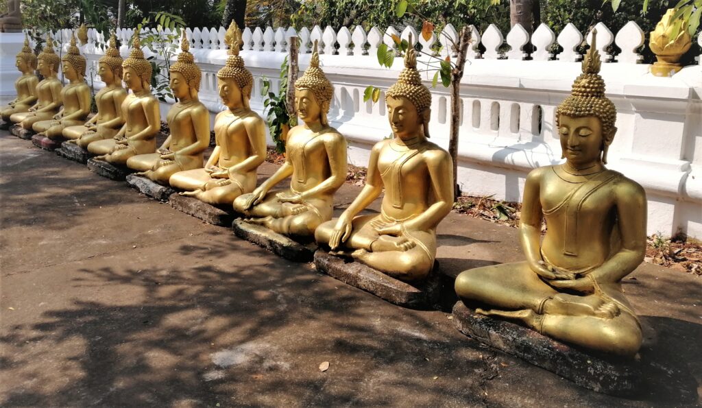 10 holy depictions sitting and meditating simultaneously in front of a long white fence on temple grounds.