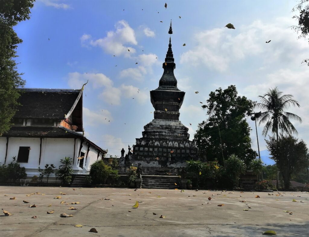 Leaves blow in the air and on pavement in front of a colossal chedi. In the post: Infinite Earth Art: Temples of Luang Prabang.