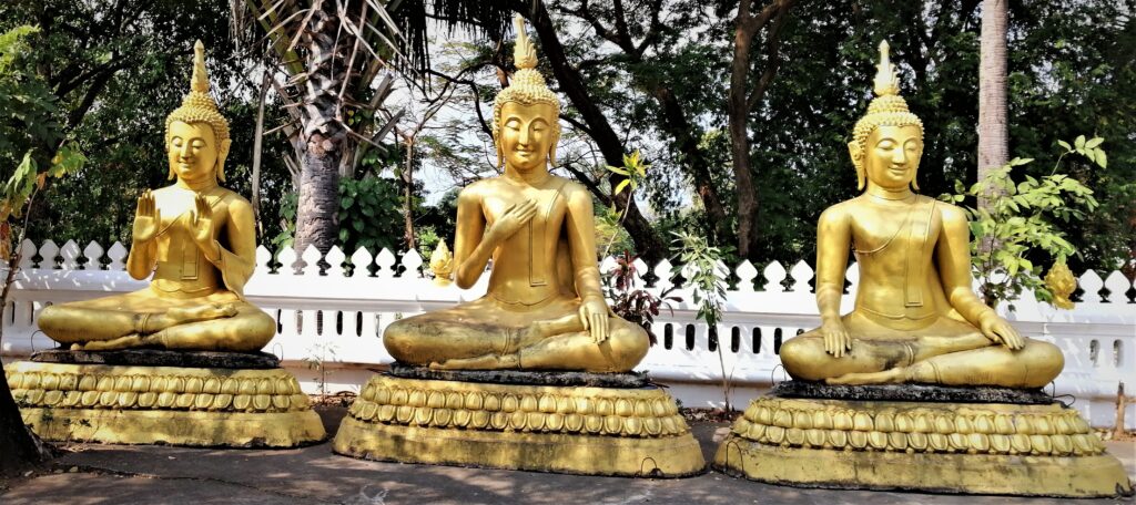 Big statues of the guatama on pedestals in front of a while fence and plants with a forest in the background. In the post: Infinite Earth Art: Temples of Luang Prabang.