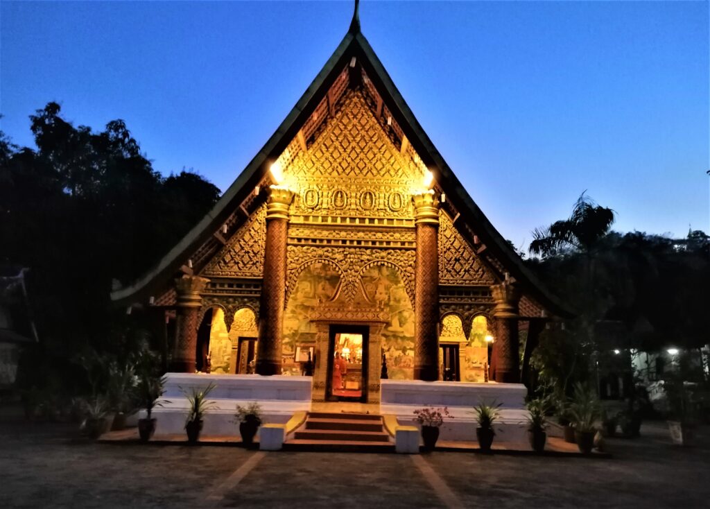 An exotic yellow temple surrounded by lush green forest under a late-afernoon, dark-blue sky. In the post: Infinite Earth Art: Temples of Luang Prabang.