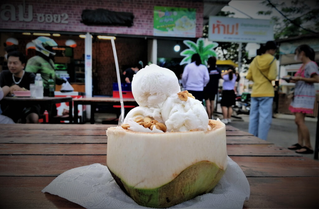 Vegan ice cream with peanuts in a coconut shell on a table.  People in the background. In post: Khon Kaen: A City of Unknown Wonders.