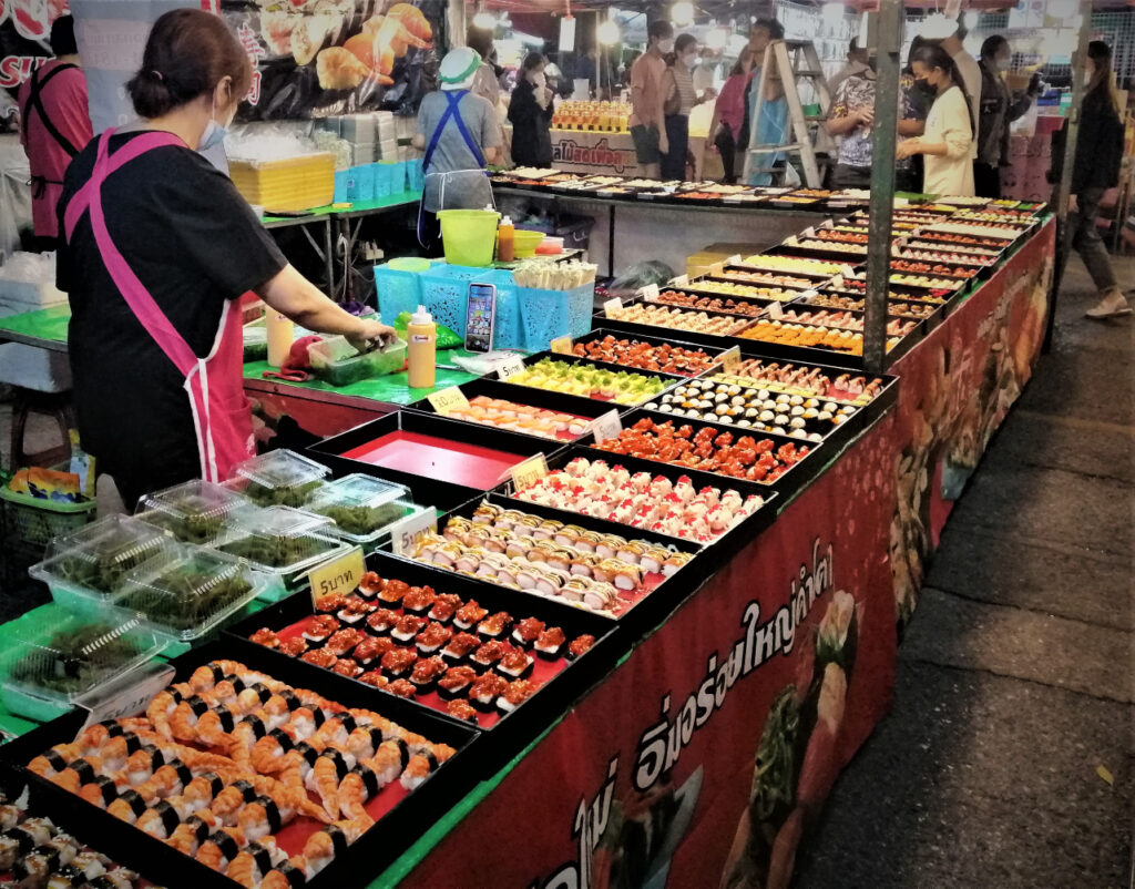 A huge sushi stall, vendor and patrons walking outside.  Thai night markets are wondrous.