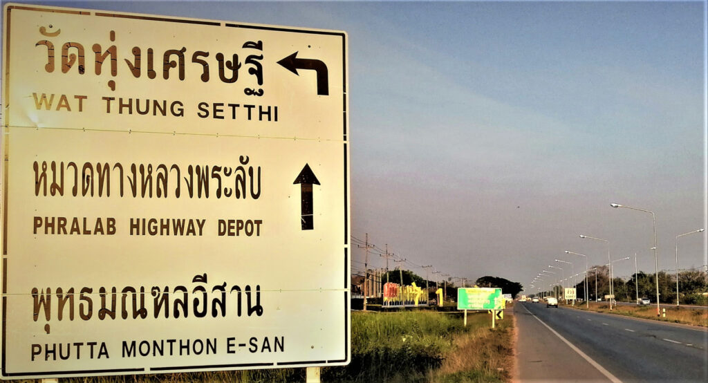 Highway sign with an arrow pointing to the wat beside a two lane highway under a wide hazy late afternoon sky. In post: Khon Kaen: A City of Unknown Wonders.