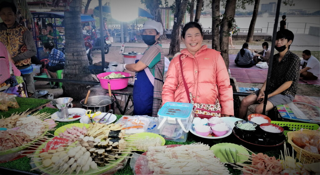 Three food hawkers smile in front of a table filled with food and a man made lake in the background. In post: Khon Kaen: A City of Unknown Wonders.