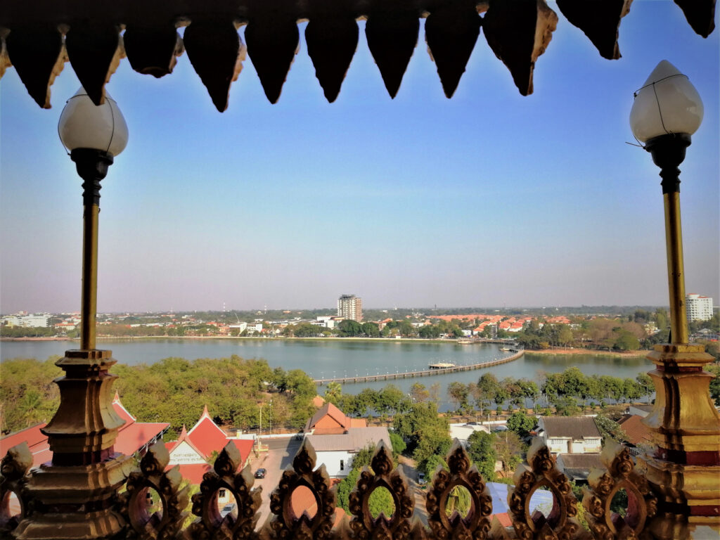 Vista of SE Asian style architecture, a manmade lake, pedestrian bridge, greenery and blue sky above.  In post: Khon Kaen: A City of Unknown Wonders