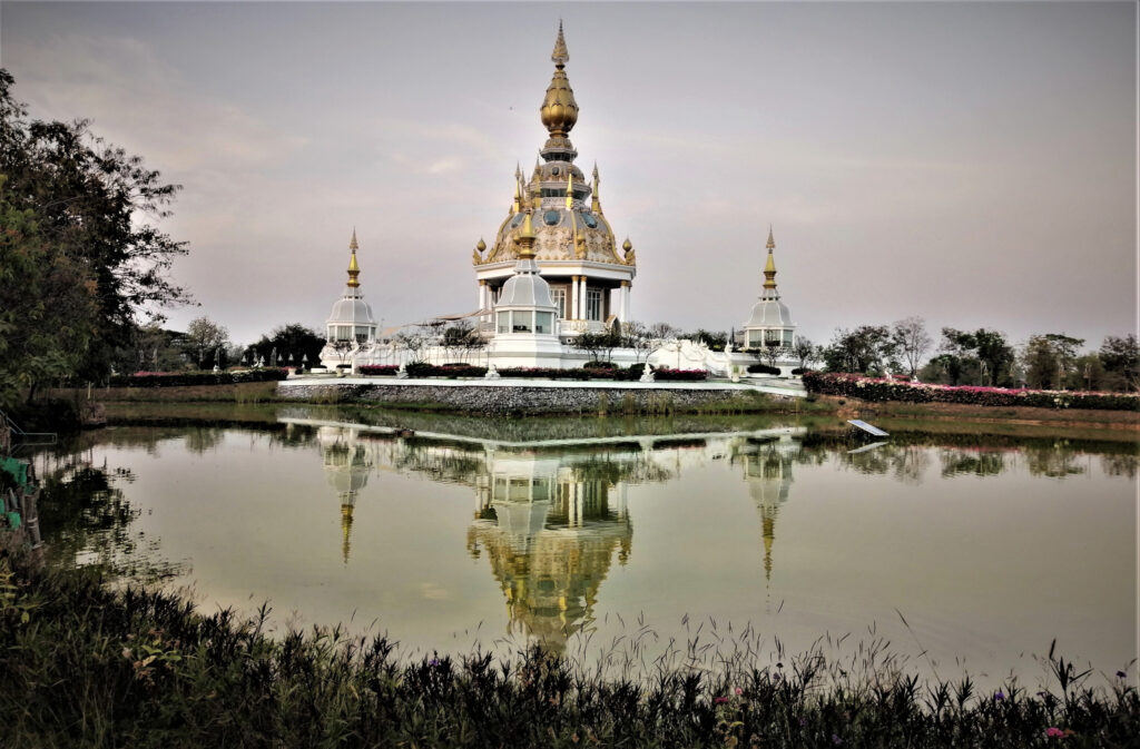 A stunning temple behind a lake with some greenery and a hazy sky at sunset.  In post: Khon Kaen: A City of Unknown Wonders.