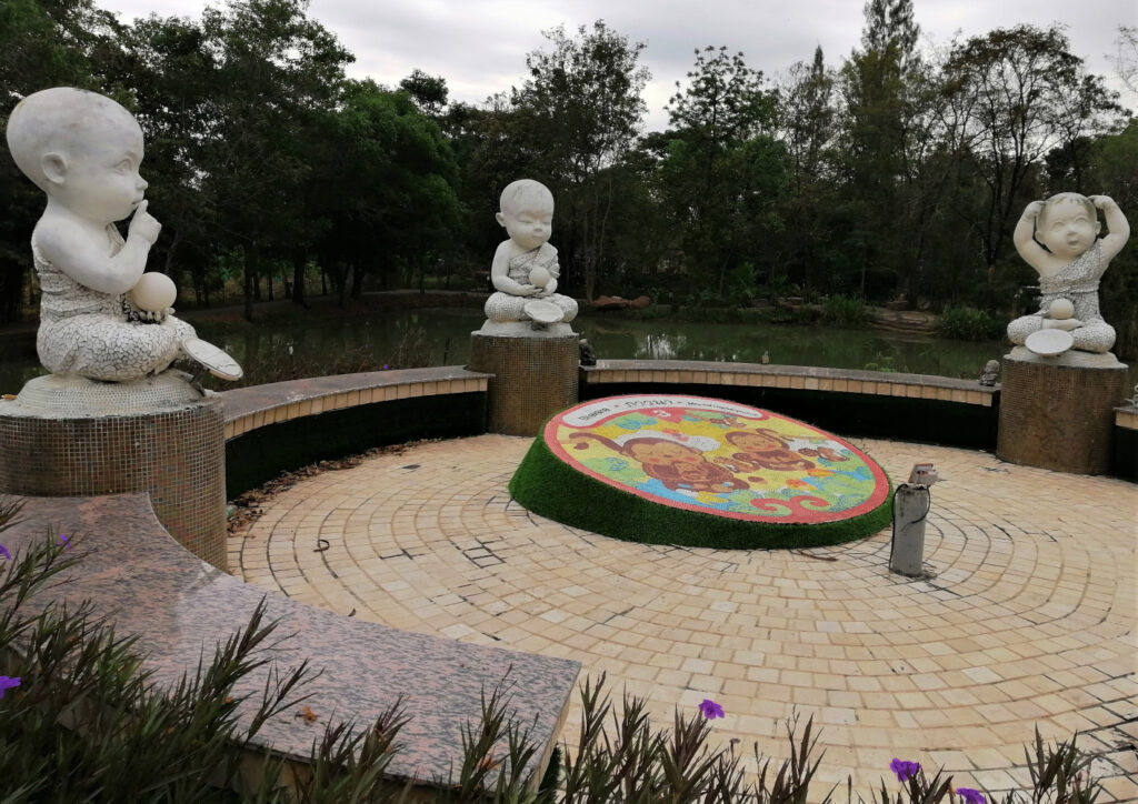 Three toddler statures over a circular display in front of a pond and forest. In post: Khon Kaen: A City of Unknown Wonders.