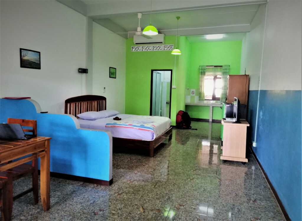 A spacious hotel room with blue, whie, and green walls.  A table, laptop, partition, queen bed, a/c and wardrobe. In post: Peaceful Pakse on the Calm Mekong. 