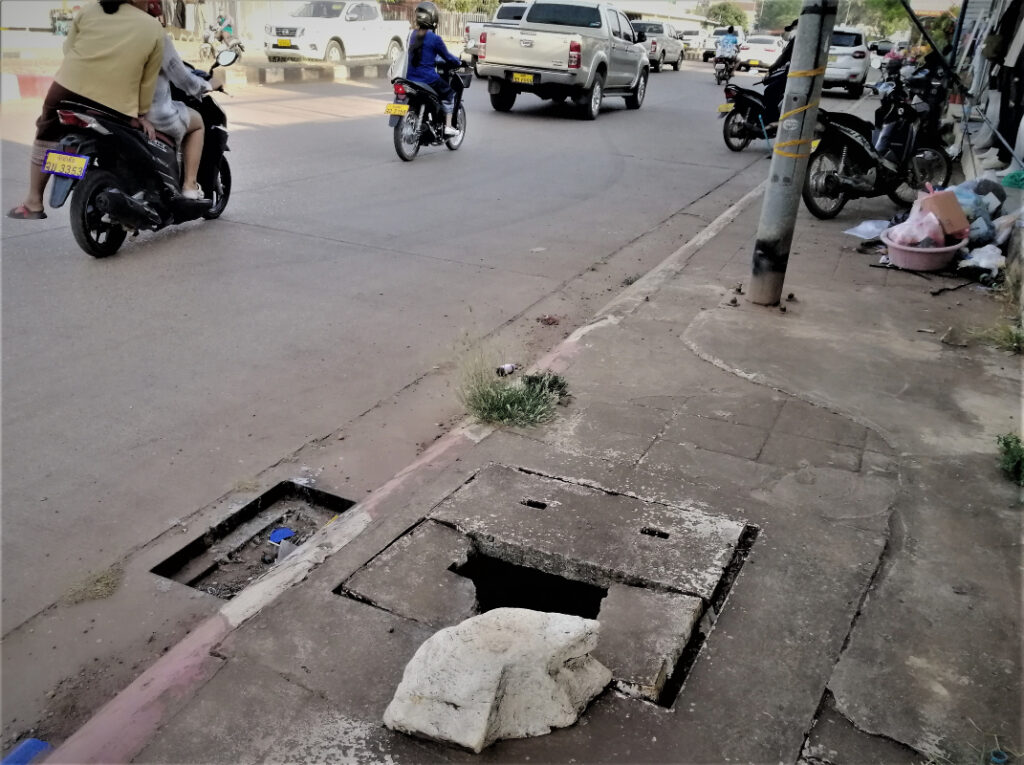A sidewalk with a dangerous hole beside a one way street busy with motorbikes and cars. In post: Peaceful Pakse on the Calm Mekong.