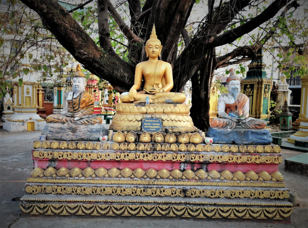 Three statues.  Buddha is centered with a disciple on either side in front of a tree on temple grounds. In post: Peaceful Pakse on the Calm Mekong.