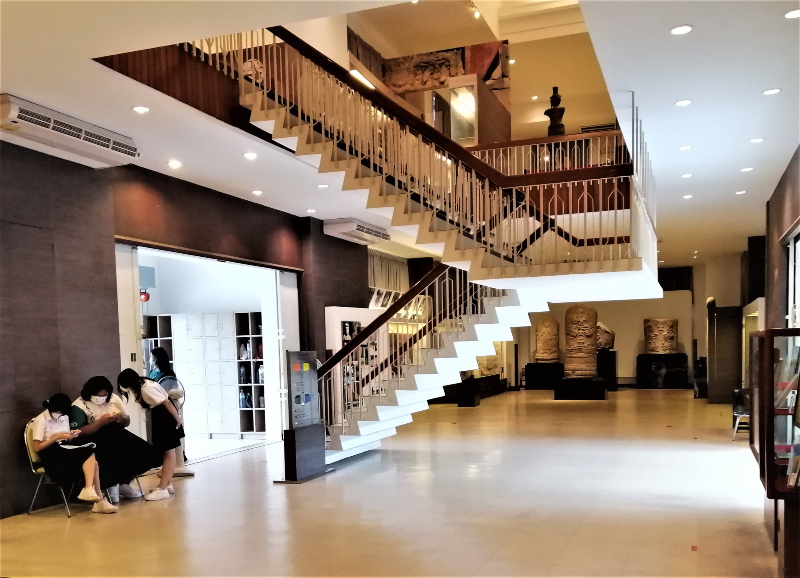 Lots of open space inside on the ground floor.  Three schoolgirls are engrossed in their and each other's phones while behind a white staircase are mammoth boundary stones on display.  In post: Visiting The Khon Kaen National Museum.