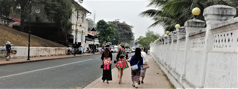 Pedestrians in traditional outfits walk on a wide widewalk along Sisavangvong Road in Luang Prabang's historic district.