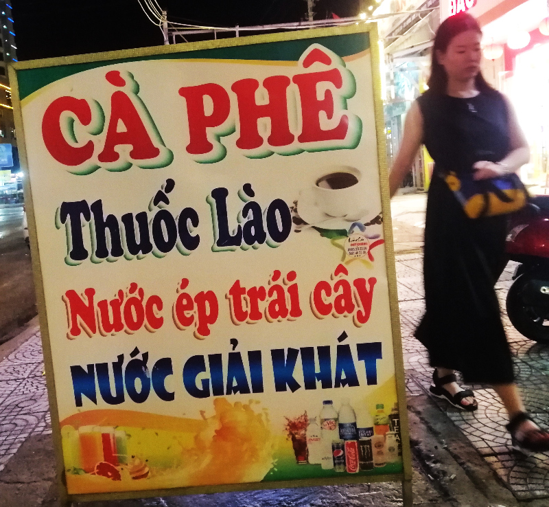 A sign in Vietnamese on a sidewalk in the evening, as a women walks past.