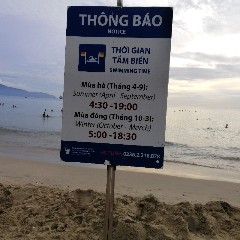 A sign that shows swimming times on and people swimming at My An Beach, Danang, central Vietnam.