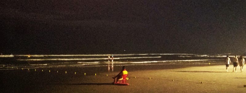 Lifeguard sits, people walk and swim, with artificial light, pre dawn, My Khe Beach, Da Nang, Vietnam.  