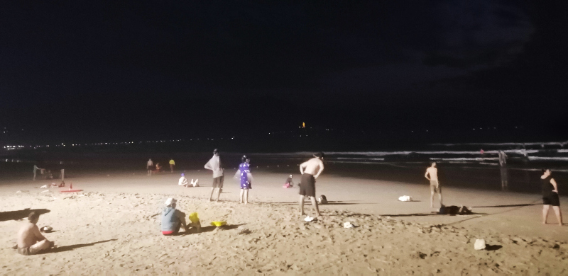 People, sand, surf and pre-dawn artificial light on My Khe Beach, Da Nang, Vietnam.