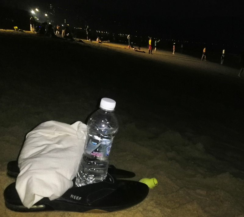 Sand, darkness, artificial light, people, sandals, T-shirt and water bottle, My Khe Beach, Da Nang, Vietnam.