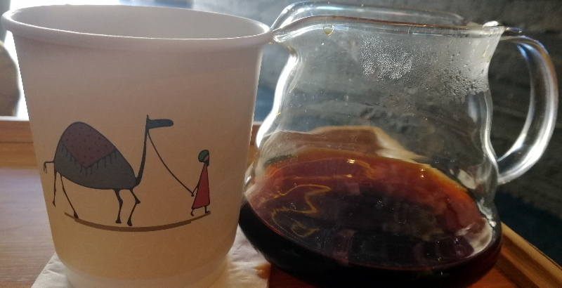 Paper cup with art: a camel and a herder, next to a pot with fresh Chemex, slow drip coffee.