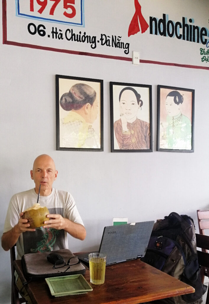 Holding a coconut with a straw, there's artwork framed on the wall behind me, inside Indochine Cafe, Danang, Vietnam.