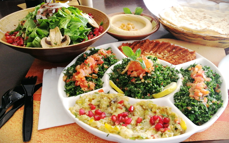 A colorful Lebanese vegan food spread consisting of arugula salad, creamy hummus, tabbouleh, babaghanoush, and mahamura.  